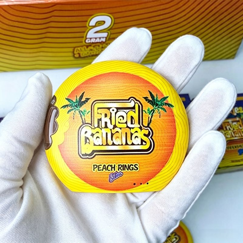 Gloved hand holding a round yellow Cookies Fried Bananas empty vape pen with strain branding.