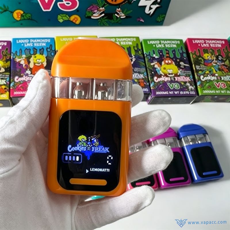 A gloved hand holding an orange 2g dual flavor disposable vape pen by Cookies x Freak Brothers V3 with packaging in background.